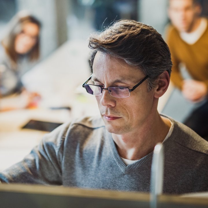 Ein Mann mit Brille arbeitet konzentriert an einem Computer, im Hintergrund sind unscharf zwei Personen zu sehen