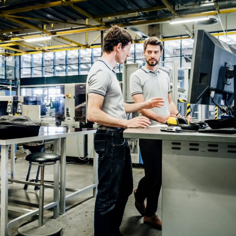 Automatisierung: Zwei Ingenieure diskutieren in einem modernen Industriebetrieb vor einem Computerbildschirm, umgeben von Maschinen und Arbeitsstationen.