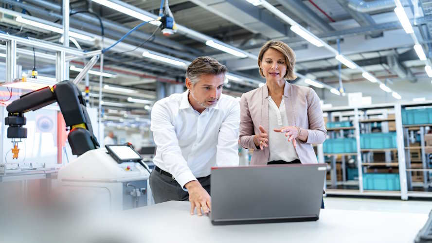 Zwei Ingenieure stehen vor Laptop in Produktionshalle und verhandeln